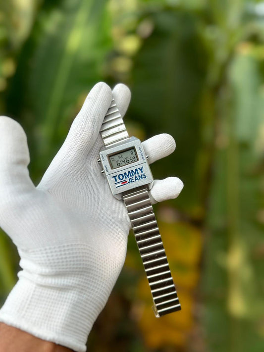 A person wearing a white glove is holding a Tommy Hilfiger watch outdoors, with plants in the background.